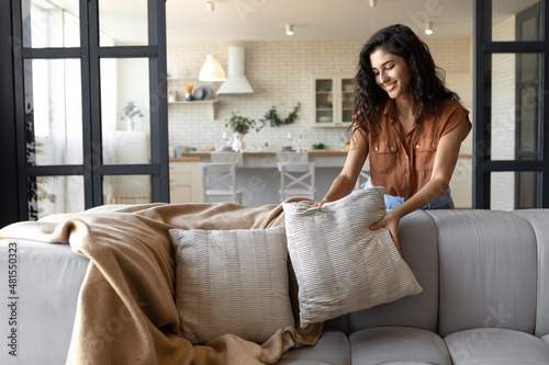 Lovely young woman putting soft pillows and plaid on comfy sofa, making her home cozy and warm, copy space