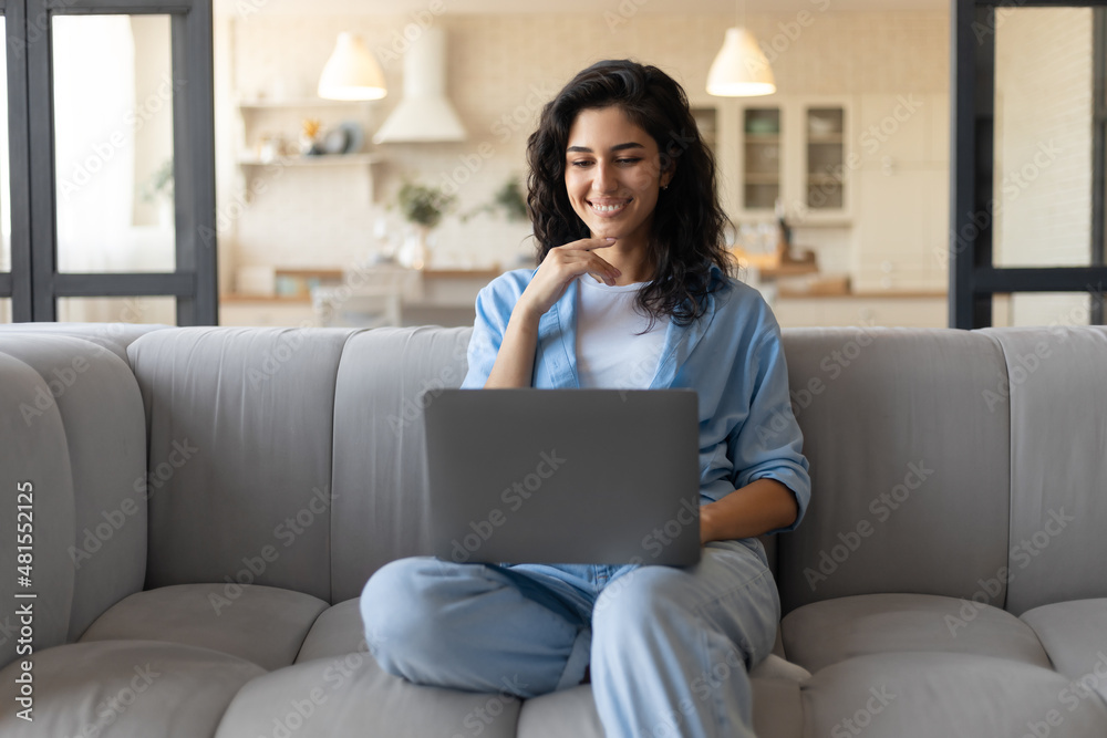 Freelancing, online job or studies. Cheerful young woman sitting on sofa with laptop, working or learning from home Freelancing, online job or studies. Cheerful young woman sitting on sofa with laptop, working or learning from home