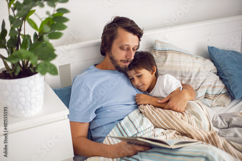 dad reads a book lying in white bed to his son child before going to bed