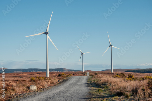 Small narrow road between fields with wing generator turbine. Warm sunny day, blue cloudy sky. Modern technology in natural environment. Source of green and renewable electricity.