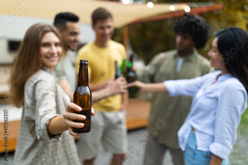 Pretty Caucasian lady with her diverse friends toasting with beer bottles, drinking alcohol near RV outdoors in autumn