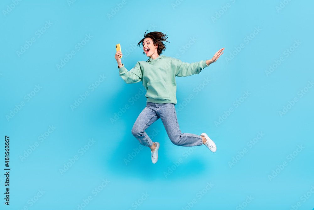 Full size photo of sweet millennial brunette lady jump look telephone wear pullover jeans footwear isolated on blue background