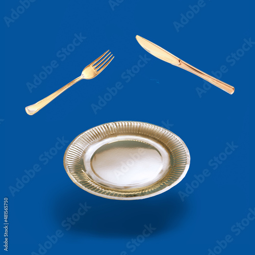 Arrangement made of levitating golden color fork, knife and plate. Dark blue background