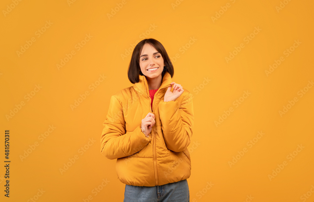 Cheerful young woman wearing and zipping up warm jacket, smiling and looking aside at free space, orange background Cheerful young woman wearing and zipping up warm jacket, smiling and looking aside at free space, orange background