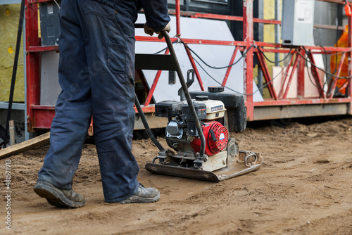 Worker use vibratory plate heavy machine compactor for construction compacting or beating sand at sidewalk. Behind a construction site when approaching a condominium residential building