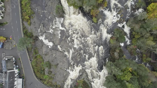 Aerial drone footage above a fast flowing water and river rapids at the Falls of Dochart in Killin, Trossachs National Park, Scotland with white water, rocks, a village and surrounded by native trees photo