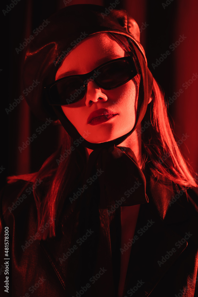 glamour woman in beret and black sunglasses posing on dark background with red light. glamour woman in beret and black sunglasses posing on dark background with red light.