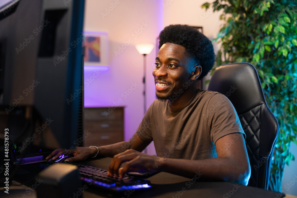 Professional male pro gamer with dark skin sits comfortably in chair leaning over computer, plays video games, smiles excitement of round, room lit with purple blue led lights