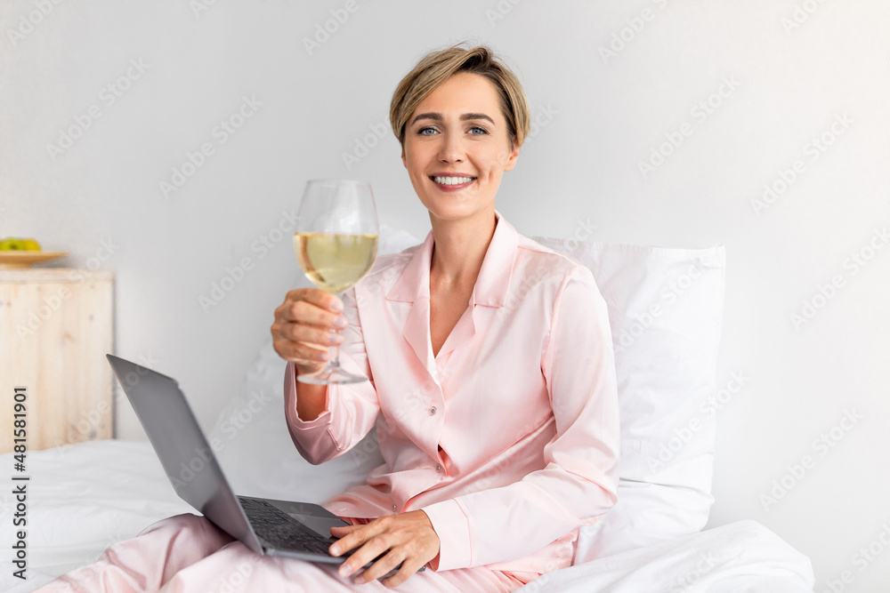 Happy woman sitting on bed using pc drinking wine Happy woman sitting on bed using pc drinking wine