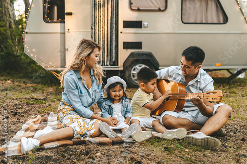 An Asian multiracial family with two children are camping in nature on a road trip. Dad plays guitar to mom daughter and son in the woods in the summer. Selective focus
