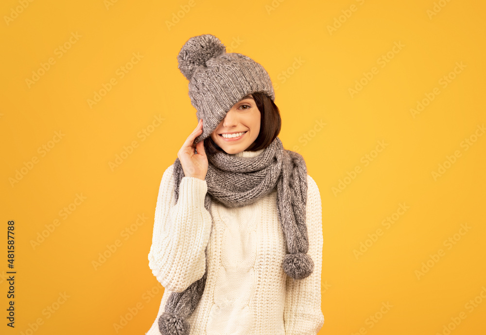 Beautiful young lady in white sweater, pulling down hat, closing one eye and smiling broadly, yellow background