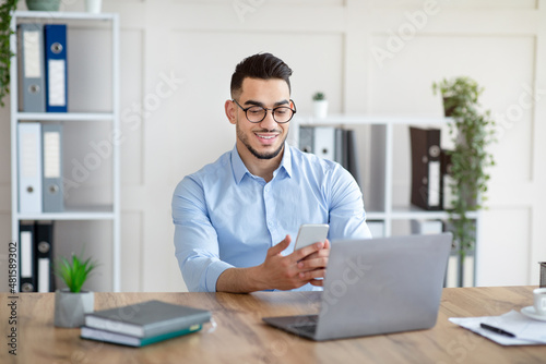 Happy young Arab man in formal wear using smartphone and laptop at workplace