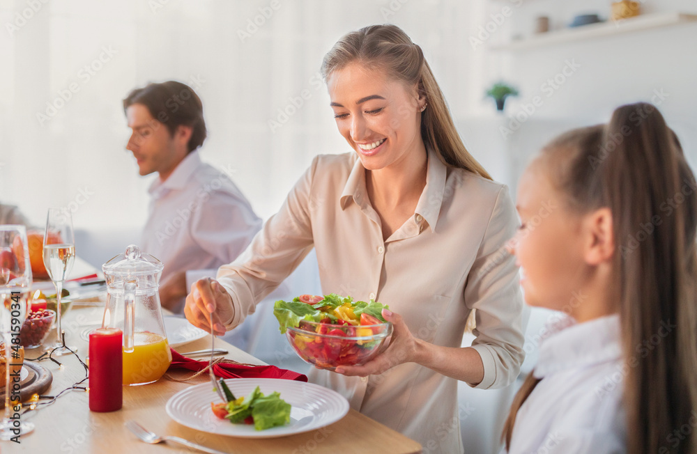 Young woman putting vegetable salad on her daughter's plate during festive family dinner at home