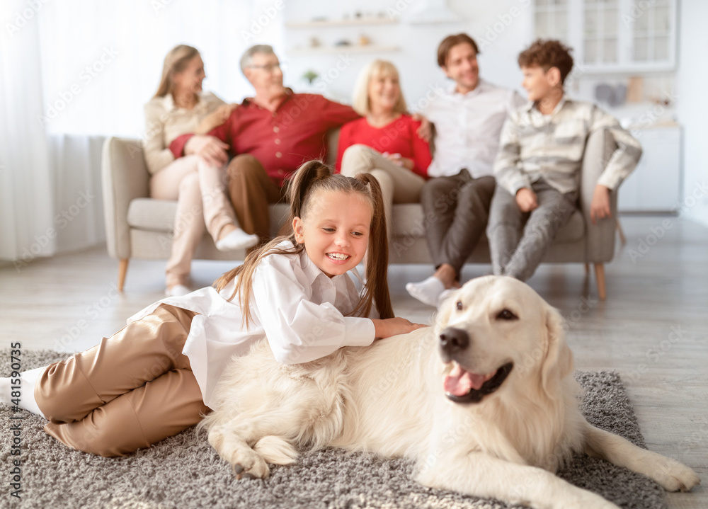 Cute little girl playing with golden retriever dog on floor in living room, her relatives resting on couch at home Cute little girl playing with golden retriever dog on floor in living room, her relatives resting on couch at home