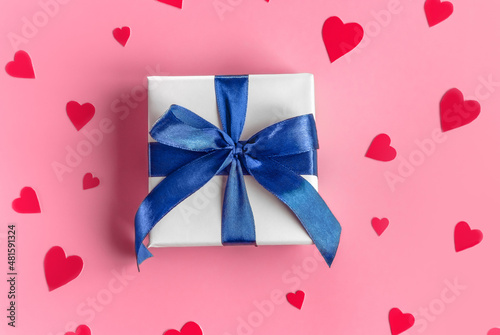Gift in white wrapping paper with a blue bow on pink background with red purple hearts. St. Valentines Day  love  tenderness  friendship  and care concept. Cozy  festive  romantic wallpaper