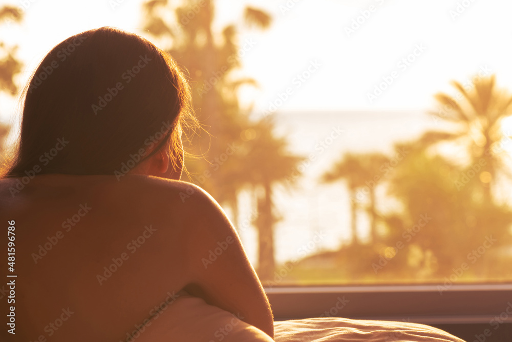 Silhouette of woman lying on pillow on bed and ready getting up early morning Female looking at sunrise. Bedroom with panoramic window and view on palm tree sea beach