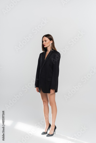 young brunette woman with naked legs, wearing black blazer and high-heeled shoes on grey.