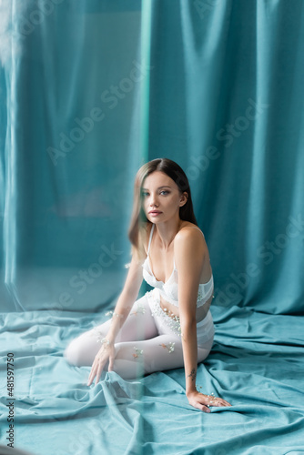 young woman in lace bra and white tights, with small flowers on body, looking at camera while sitting on green drapery.