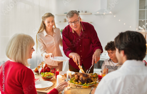 Happy elderly man cutting festive roasted turkey, celebrating Thanksgiving or Christmas with his family at home