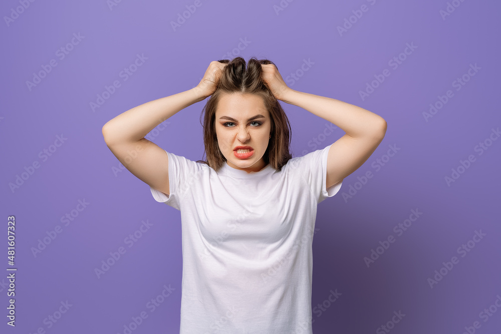 Shocked concerned young woman, hold hands on head, look angry and furious facing difficulties, standing in blank white t shirt over purple background Shocked concerned young woman, hold hands on head, look angry and furious facing difficulties, standing in blank white t shirt over purple background