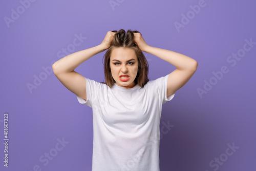 Shocked concerned young woman, hold hands on head, look angry and furious facing difficulties, standing in blank white t shirt over purple background