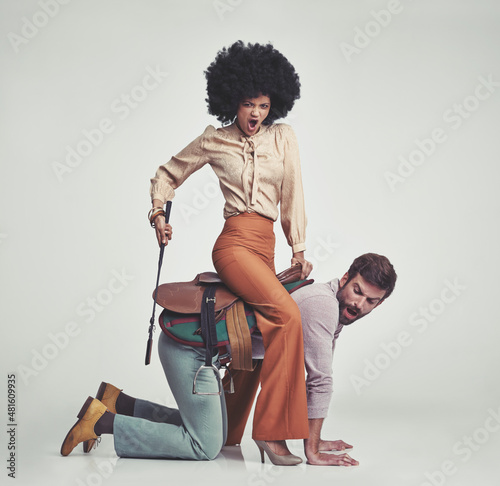 Get moving. A studio shot of an attractive woman in 70s wear riding a handsome man wearing a saddle while using a riding crop.