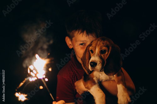 Closeup boy and dog (beagle puppy) having fun with bengal lights at night event (4th of July). Happy boy holding burning fireworks outside
