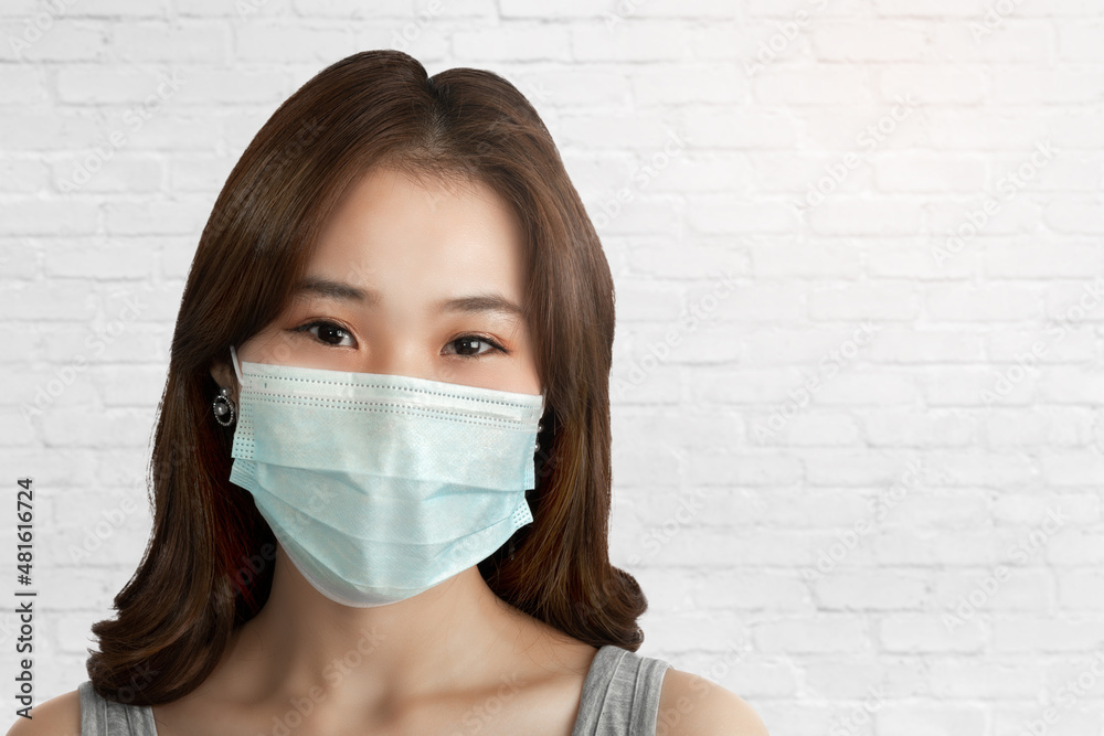 Close-up of a young Asian woman with a smile under a sanitary face mask against coronavirus or COVID-19 over an old white brick wall. Virus and illness protection, sanitizer in a public crowded place Close-up of a young Asian woman with a smile under a sanitary face mask against coronavirus or COVID-19 over an old white brick wall. Virus and illness protection, sanitizer in a public crowded place