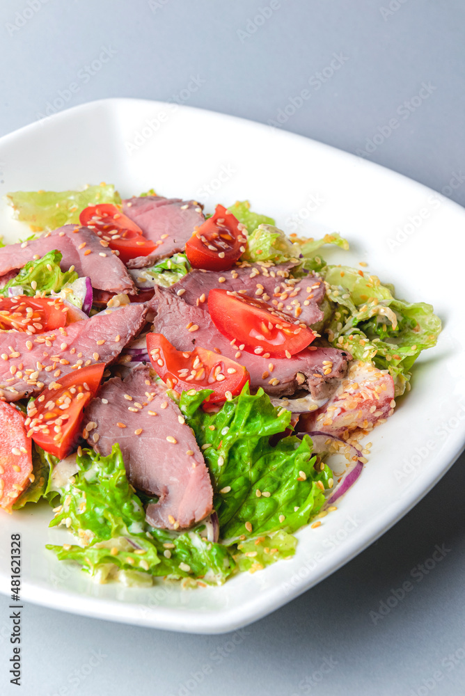 Healthy green salad with meat, tomato and lettuce served in a square white plate over grey or gray background. Healthy green salad with meat, tomato and lettuce served in a square white plate over grey or gray background.