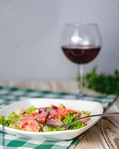 Healthy green salad with meat  tomato and lettuce served in a square white plate with a glass of red wine.