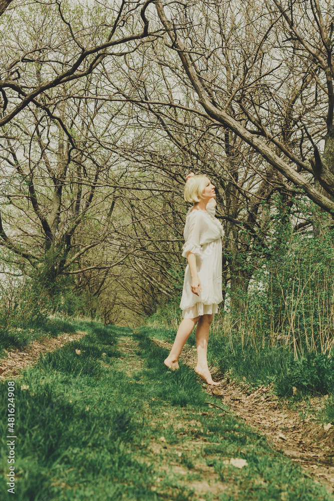 Beautiful young woman in white chiffon dress with short blonde haircut walking along barefoot the path and looking at the trees Beautiful young woman in white chiffon dress with short blonde haircut walking along barefoot the path and looking at the trees