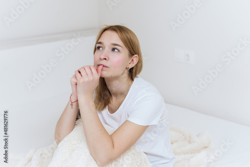 A young woman sits in bed, worries and bites her nails. Worried casual woman sitting on her bed in bedroom