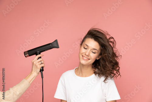 Portrait of curly woman with hairdryer and long cord on pink background