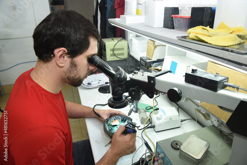 A young man in the workshop examines the microcircuit of an electrical device through a microscope, performing electrical soldering of equipment