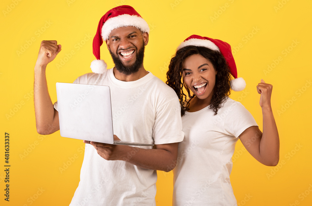 Christmas offer. Overjoyed black spouses in santa hats celebrating success with laptop over yellow background Christmas offer. Overjoyed black spouses in santa hats celebrating success with laptop over yellow background
