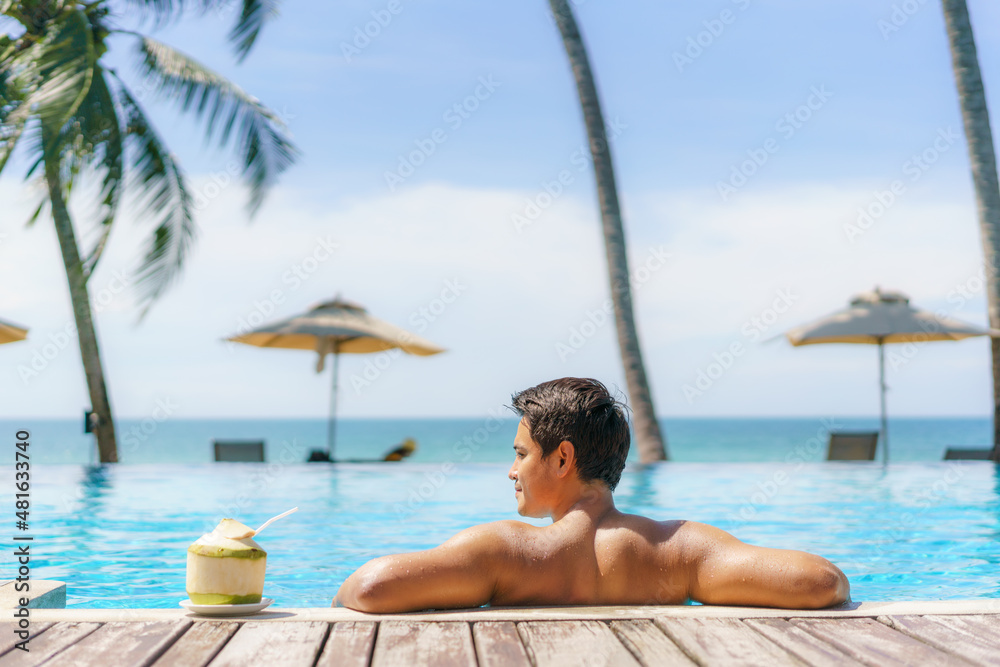 Summer travel vacation concept, Traveler asian man with coconut relax in luxury infinity pool hotel resort with sea beach and palm tree background