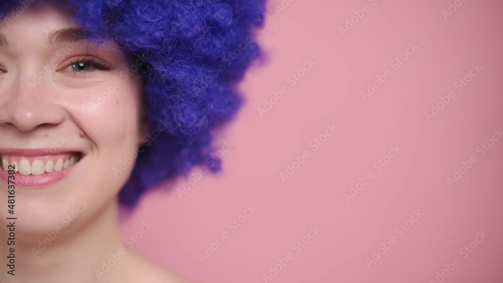 Young woman in fancy blue wig opens her eyes and smiles. Close-up portrait on pink background. Cropped shot. Young woman in fancy blue wig opens her eyes and smiles. Close-up portrait on pink background. Cropped shot.