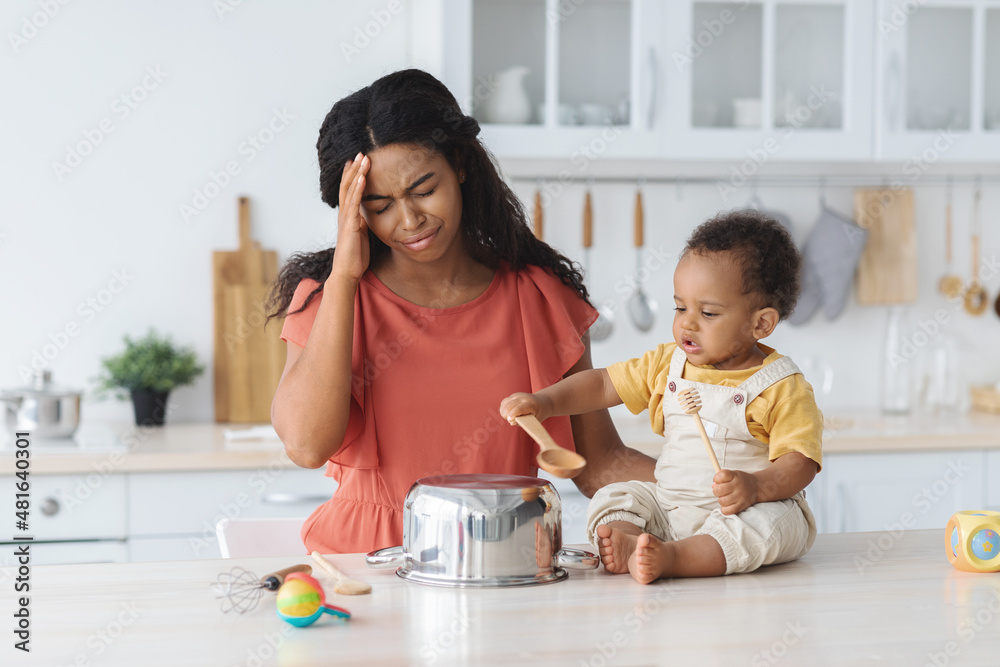Black mother suffering headache while her infant baby making noise in kitchen Black mother suffering headache while her infant baby making noise in kitchen