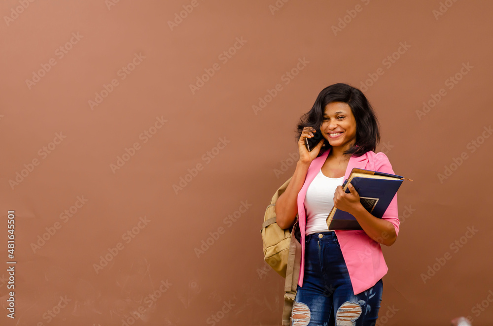 Young black African female college student standing on a grey background holding a backpack and schools book smiling making a phone call read for lectures Young black African female college student standing on a grey background holding a backpack and schools book smiling making a phone call read for lectures