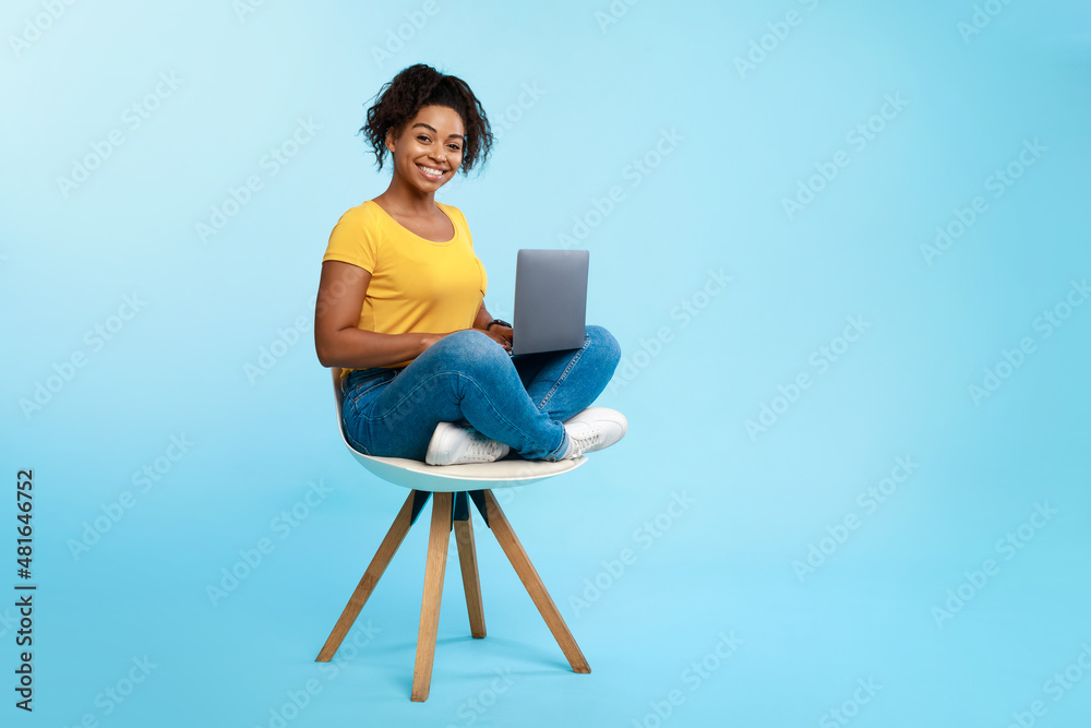 Happy young Afro lady sitting on chair, using laptop, studying online or having business meeting on blue background Happy young Afro lady sitting on chair, using laptop, studying online or having business meeting on blue background
