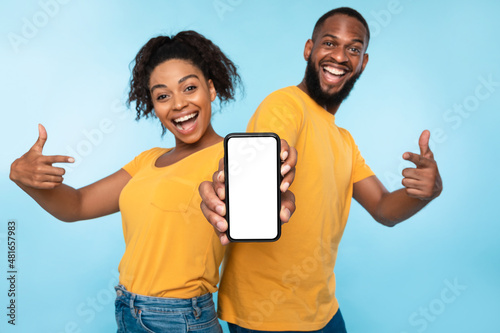 Excited Afro couple pointing at cellphone with blank screen, recommending new app on blue background, mockup for website