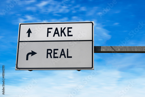 Road sign with words fake and real. White two street signs with arrow on metal pole on blue sky background.