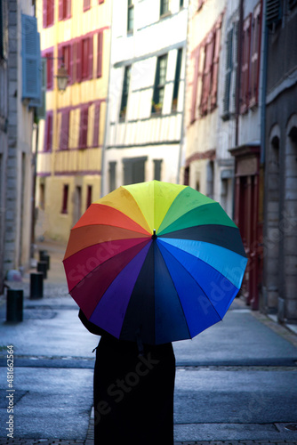 Una persona con ropa negra y un paraguas con los colores del arcoiris de espaldas paseando por una calle de un pueblo frances © Safi