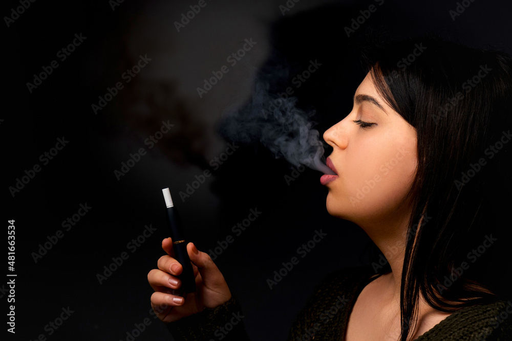 Fototapeta premium A young woman smokes a vape and lets out smoke on a dark background, Vaping concept with place for text.