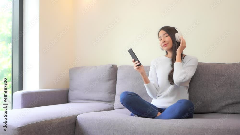 A young woman sitting cross-legged on the couch and wearing headphones sways to the music streaming through her smartphone. A young woman sitting cross-legged on the couch and wearing headphones sways to the music streaming through her smartphone.