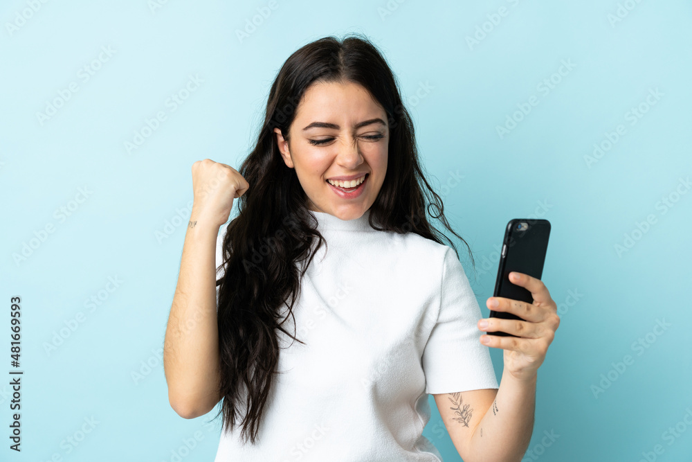 Young woman using mobile phone isolated on blue background celebrating a victory Young woman using mobile phone isolated on blue background celebrating a victory