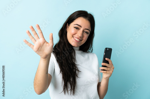 Young woman using mobile phone isolated on blue background saluting with hand with happy expression