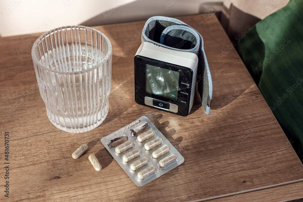 Obraz premium Pills capsules, glass of water and digital Blood Pressure Monitor on wooden background