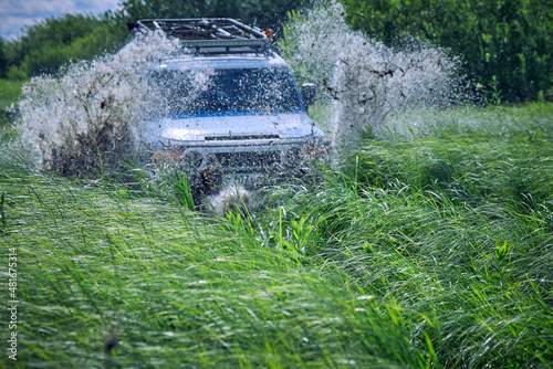 Off-road car crashed into a puddle and raised mud splashes. A four-wheel drive sports car is racing among the green grass. Off-road 4x4 SUV in blue color. The best off-road vehicles