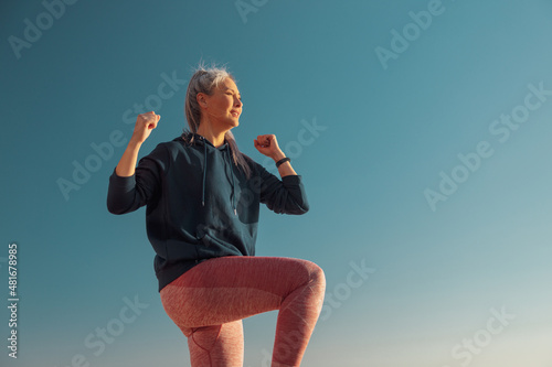 Active female in pink leggins and blue hoodie exercising , lifting one knee up and looking ahead on sunny day photo